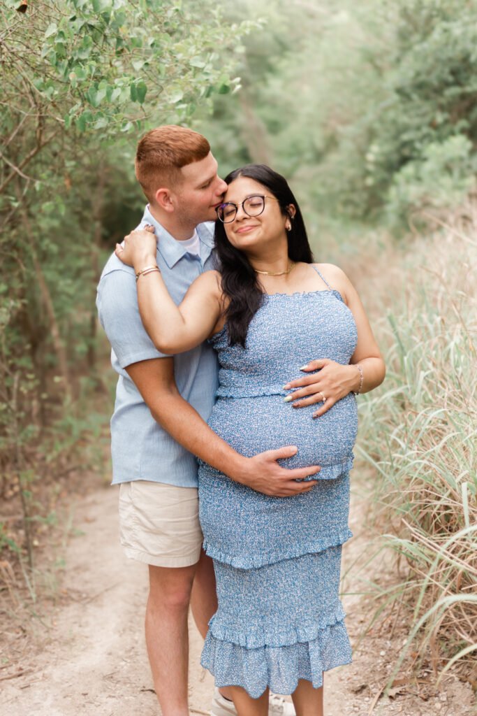 Wife holds onto husband while he lays his hand on her stomach for maternity session